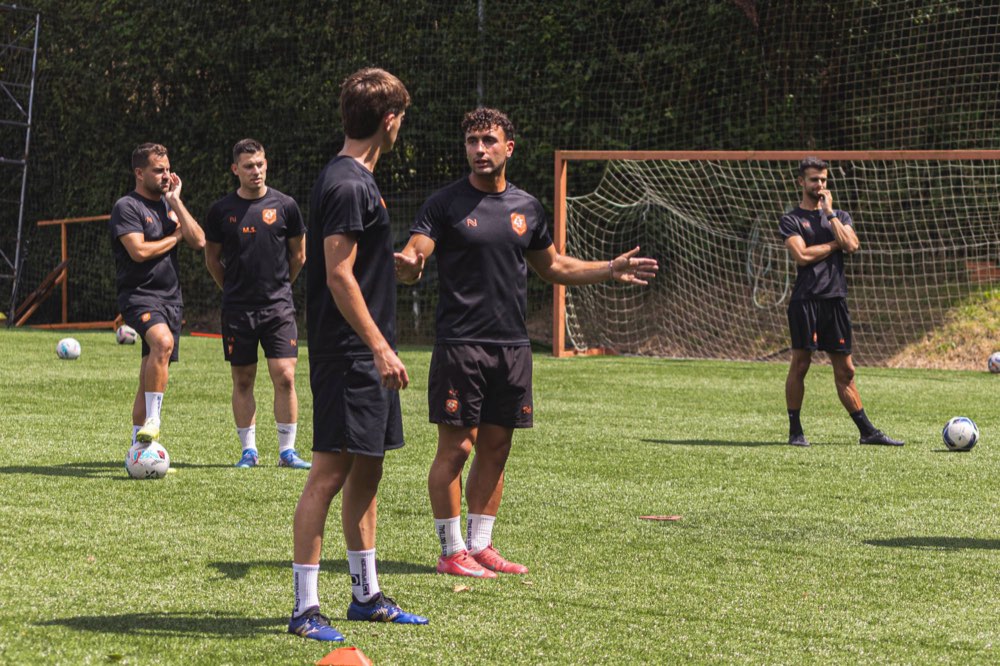 Un entrenador de L4F PRO corrige a un futbolista en el campo de la academia mientras el equipo técnico observa